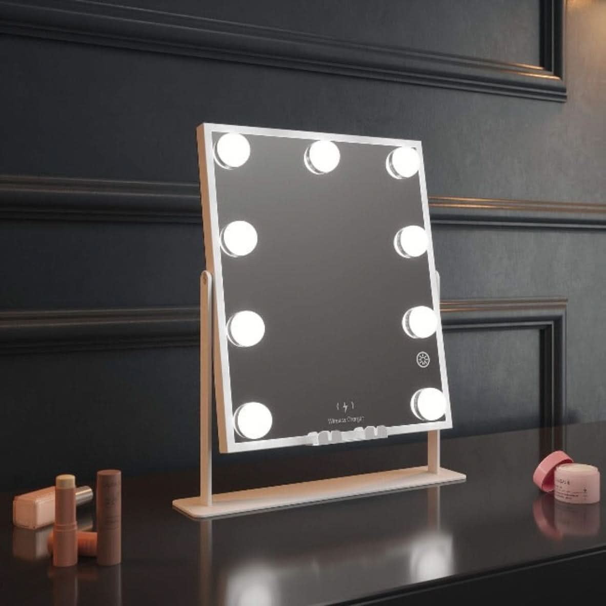 The Renée Bluetooth Hollywood Mirror 30x25cm, featuring eight LED bulbs, sits on a dark counter among makeup and skincare items, set against a paneled wall.
